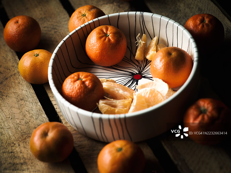 一个陶瓷碗里和周围放着完整和剥皮的橘子，摆放在一张旧木桌上。图片素材