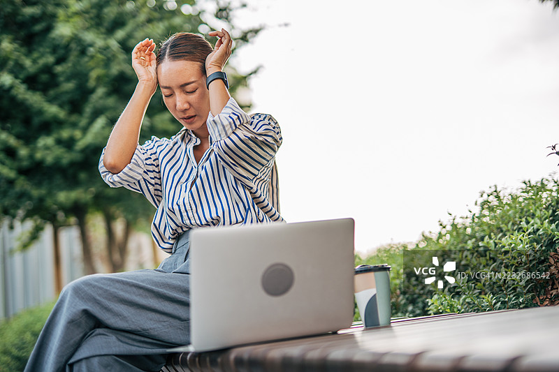 在户外用笔记本电脑工作时，感到压力的女商人捂着头。图片素材
