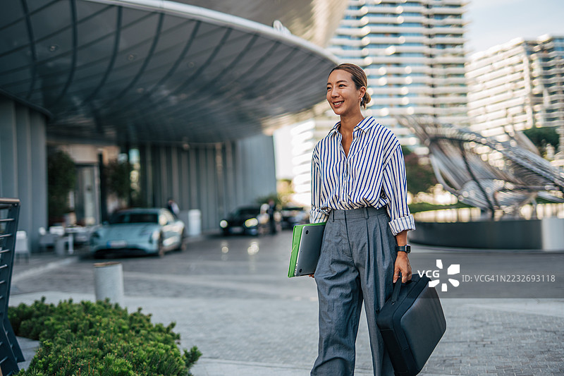 微笑的女商人在办公楼外走着，手里拿着笔记本电脑和包。图片素材