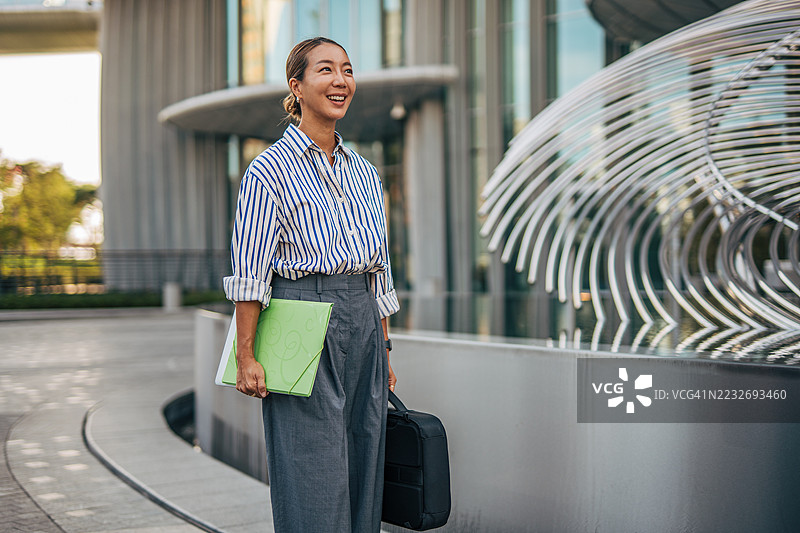 微笑的女商人在办公楼外走着，手里拿着文件夹和公文包图片素材