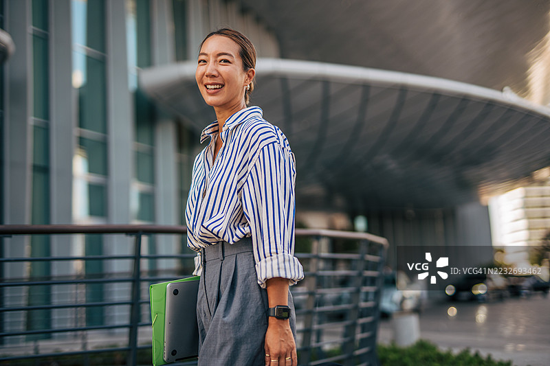 微笑的女商人在办公楼外走着，手里拿着笔记本电脑图片素材