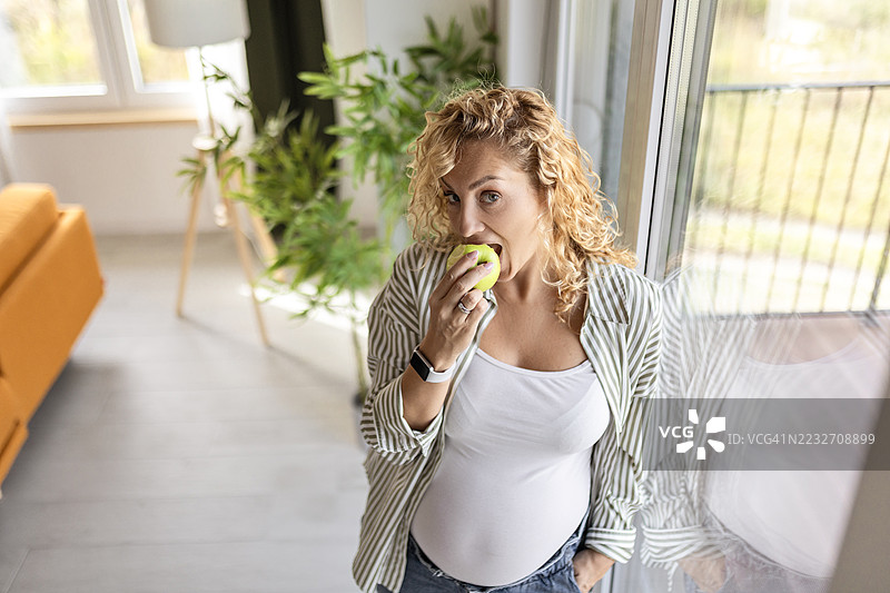 美丽的孕妇在家里吃苹果并看着镜头图片素材