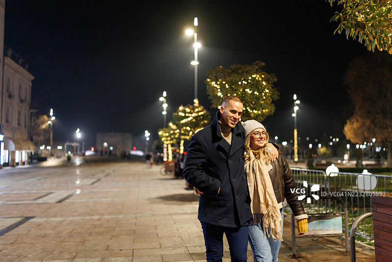 快乐的年轻情侣在装饰有圣诞灯饰的城市中享受傍晚散步。图片素材