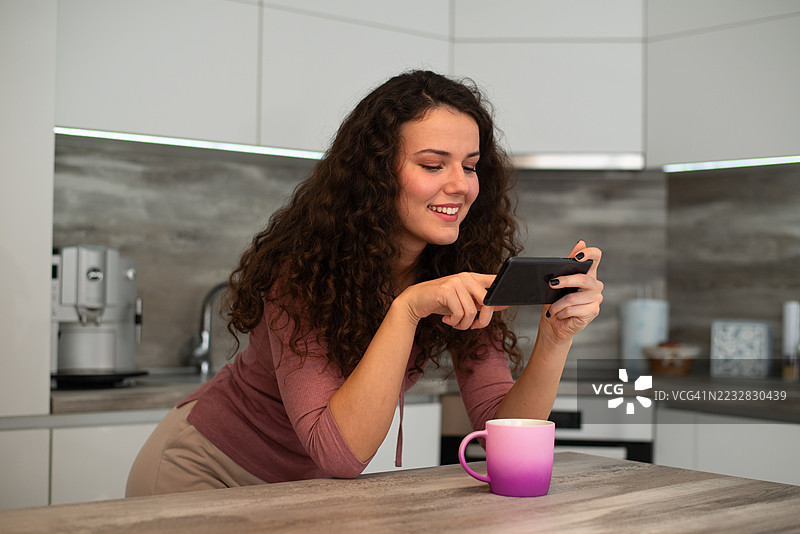 年轻快乐的女人在厨房喝咖啡并使用手机图片素材