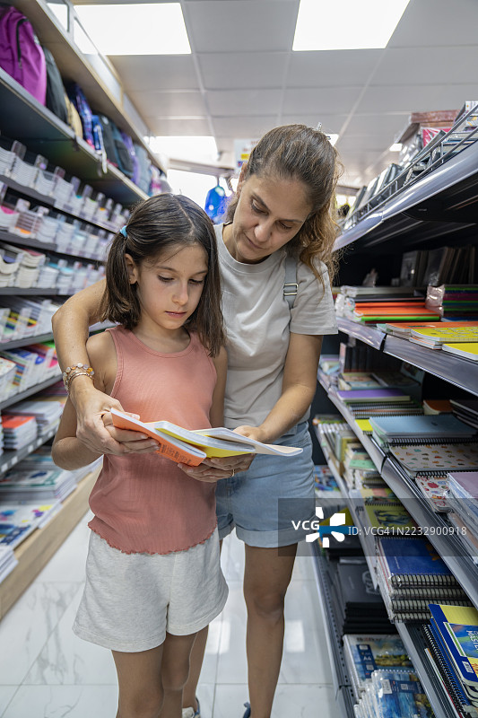 年轻母亲和女儿在书店里寻找笔记本和书籍。返校购物概念。为上学购物。家长在考虑买哪个笔记本。图片素材