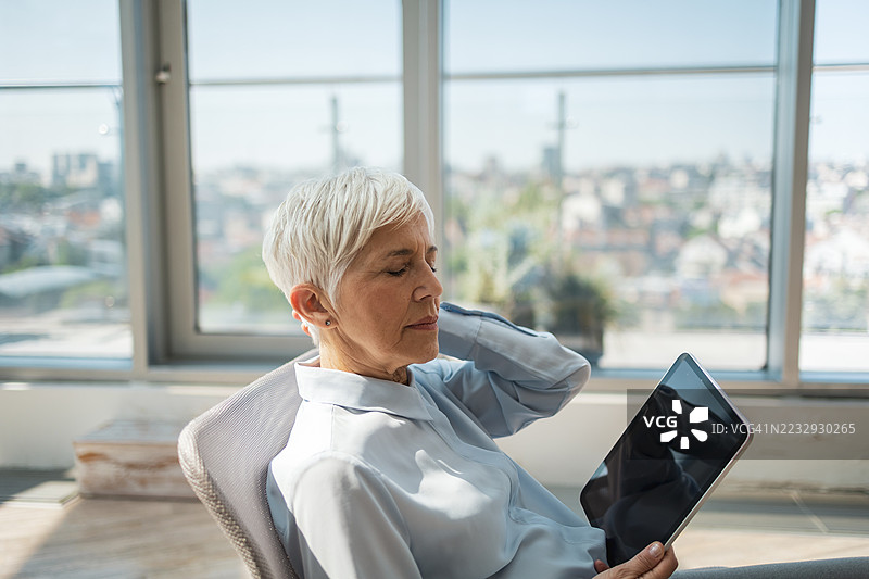 一位老年女性在现代阳光明媚的办公室里放松，手中拿着平板电脑。图片素材