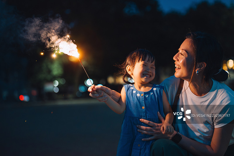 快乐的年轻亚洲母亲和小女儿在夜晚的公园里玩烟花。家庭享受着与烟花棒一起的美好时光。母女时刻。家庭生活方式。家庭的爱与团聚。图片素材