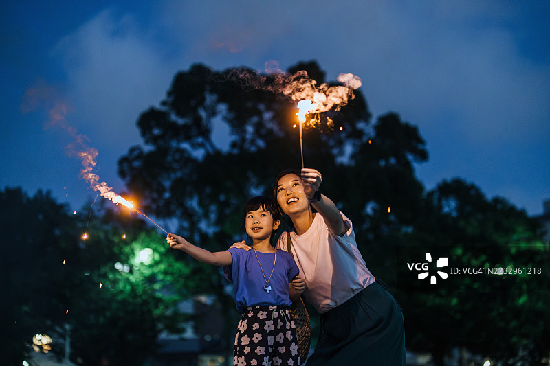 快乐的年轻亚洲母亲和女儿在夜晚的公园里玩烟花。家庭享受着与烟花棒一起的美好时光。母女时刻。家庭生活方式。家庭的爱与团聚。图片素材