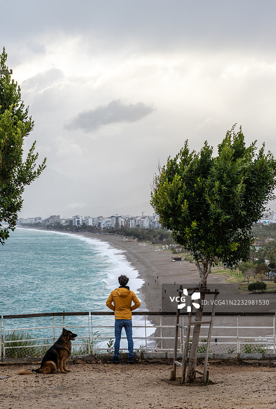 穿着黄色夹克的年轻人与他的狗站在栏杆旁，凝视着孔亚尔提海滩。图片素材
