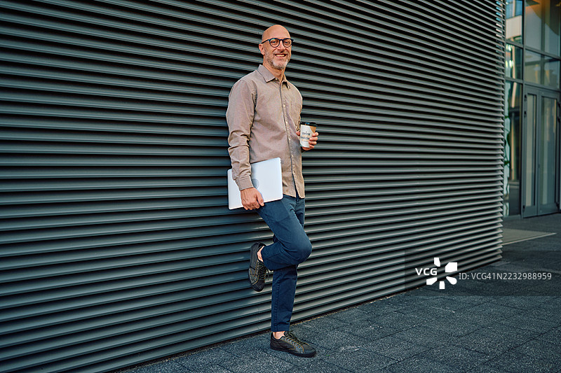 微笑的商人手持笔记本电脑和外卖咖啡，靠在墙上图片素材