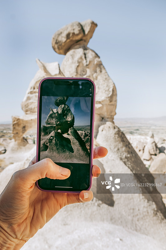 女性手持智能手机拍摄一处形似骆驼的岩石 formations。聚焦于手机屏幕。移动摄影和旅游体验概念。图片素材
