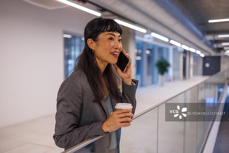 现代办公室里，女商人一边打电话一边喝咖啡。图片素材