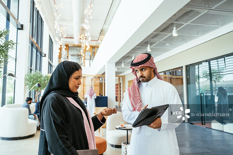 多元化的阿拉伯商人穿着头巾和长袍，在现代明亮的办公室中一起使用数字平板进行简报。图片素材