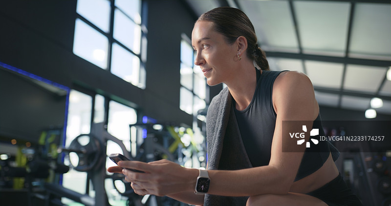 女性在健身房休息时使用手机发短信，反思健身或使用健康应用。此人正在训练俱乐部中滚动手机，思考、联系并休息，进行锻炼计划。图片素材