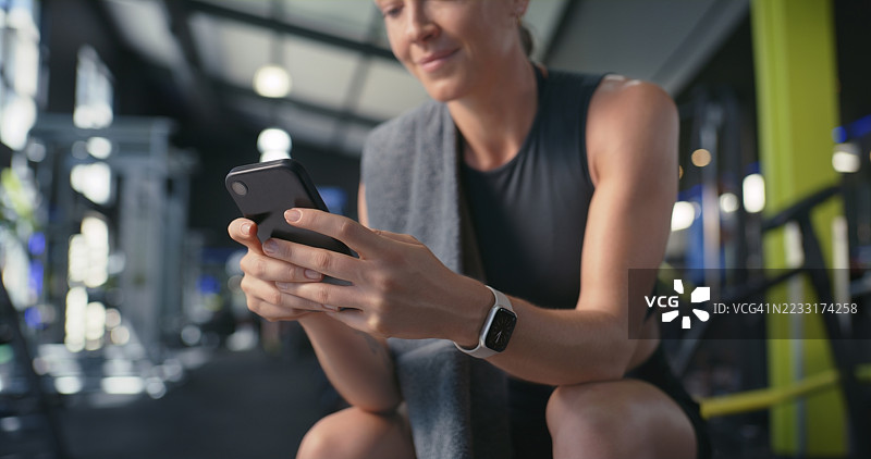 在健身房休息时，手握手机发短信，查看统计数据、结果和健康应用程序。此人正在训练俱乐部使用智能手机滚动聊天、联系或休息，查看锻炼计划。图片素材