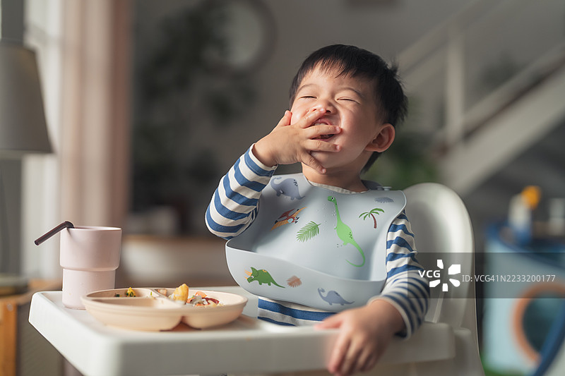 幽默的亚洲幼儿男孩在吃晚餐时试图闭上眼睛，男孩坐在家中客厅的高脚椅上。图片素材