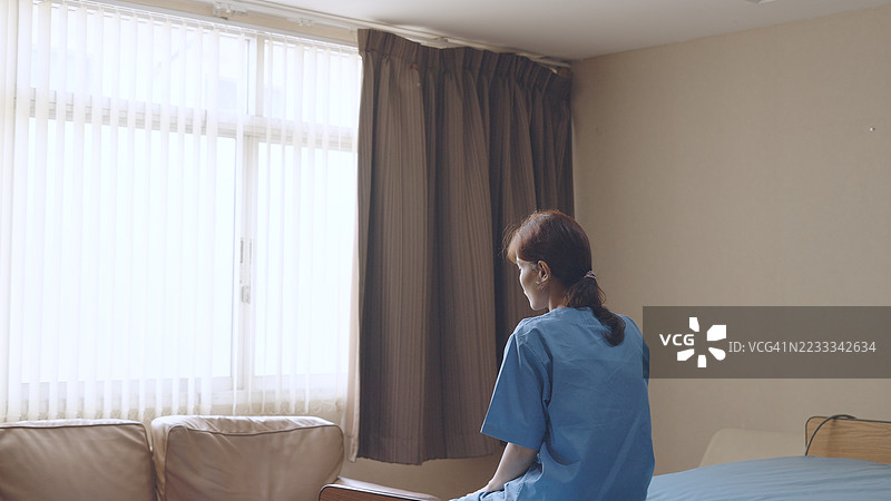 年轻女性穿着蓝色病人制服，独自坐在医院私人病房的床上，望着窗外，表达对检查结果的担忧。悲伤的病人，思考着压力和焦虑，伴有抑郁症状。图片素材