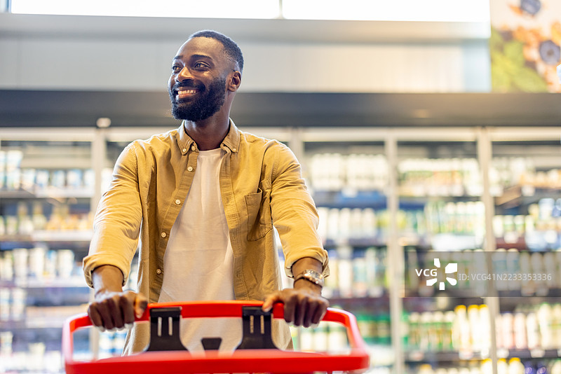 快乐的男人在杂货店购物，推着红色购物车穿过过道图片素材