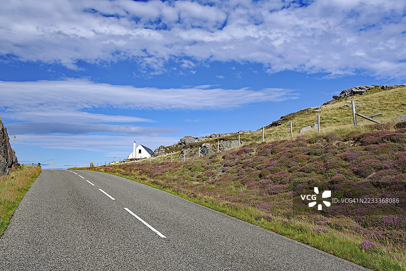孤独的道路穿过丘陵风景，背景是一座白色的小屋，位于苏格兰赫布里底群岛的刘易斯岛。图片素材