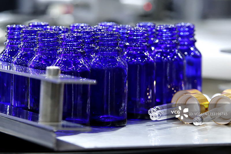 一组小巧、鲜艳的蓝色玻璃瓶，配有螺旋盖，整齐地排列在不锈钢表面上，旁边有金色瓶盖的滴管，象征着药品、化妆品或精油包装。图片素材