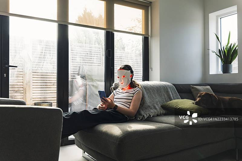 一个少女在家沙发上放松时使用红色LED面膜（红光疗法），同时使用她的智能手机。图片素材