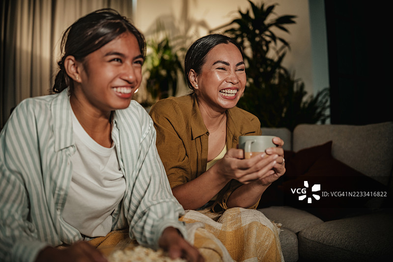 母女俩在家享受温馨的夜晚，一边分享欢笑和零食，一边观看她们最喜欢的节目。图片素材