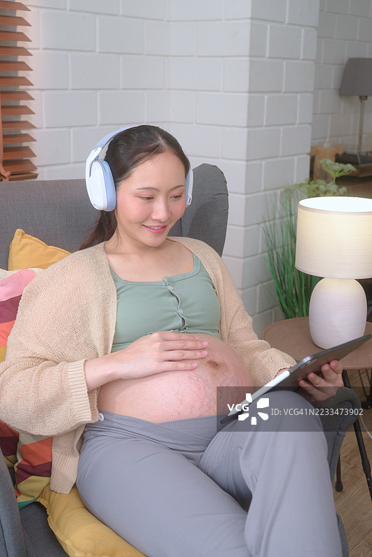 亚洲孕妇戴着耳机，使用平板电脑搜索怀孕信息图片素材