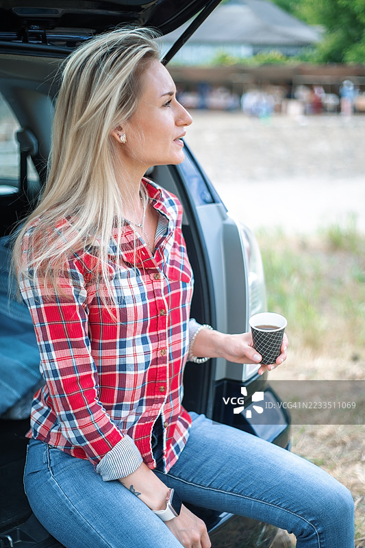年轻的金发女性坐在汽车后备箱里喝咖啡图片素材