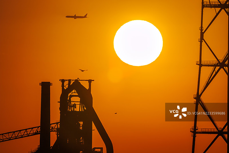 第一缕阳光与一架客机和鸟的剪影。它即将降落在附近的羽田机场。第一缕阳光从运河对岸的烟囱和电厂、钢铁厂上升。横滨，神奈川。图片素材