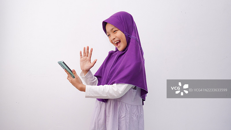 穆斯林小女孩在白色背景下使用智能手机进行视频通话，摄影棚拍摄概念图片素材