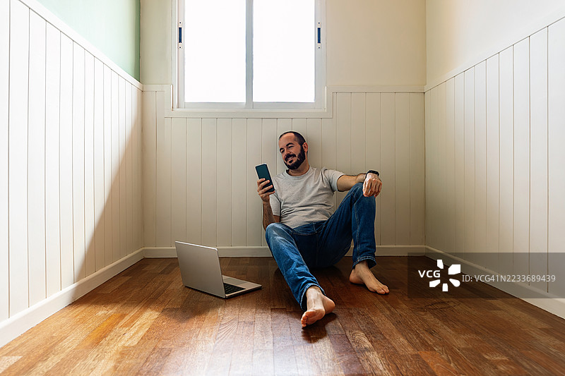 年轻快乐的男子在空房间的地板上使用智能手机，同时在笔记本电脑上工作图片素材