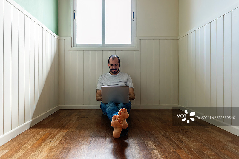 中年男子坐在空荡荡的家居房间地板上使用笔记本电脑。图片素材