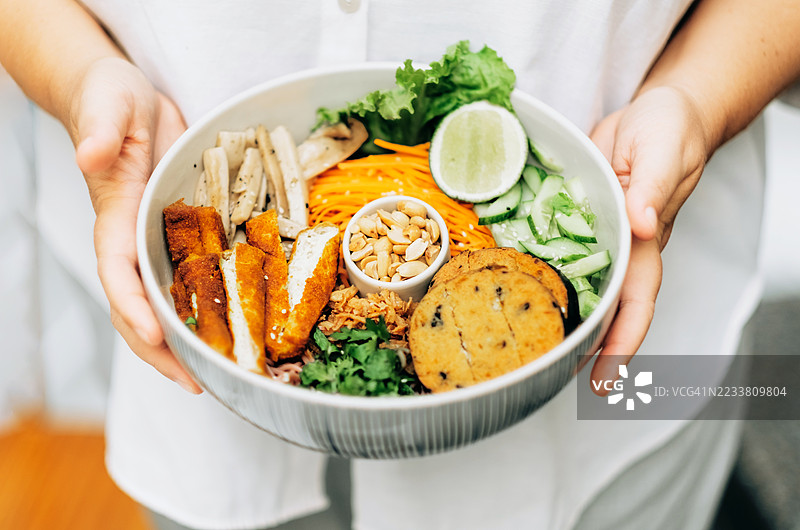 女人手持一碗混合的素食食品，包括豆腐、黄瓜、 shredded 胡萝卜、生菜、花生、青柠和素食饼。图片素材