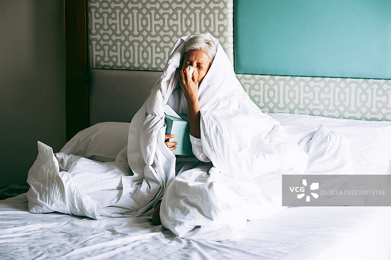 短发灰色的女人坐在床上，裹着毯子，用纸巾擤鼻子。手里拿着杯子和书。感冒或流感恢复的概念。图片素材