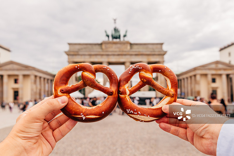 朋友们在德国柏林的勃兰登堡门附近手持两个椒盐卷饼，从个人视角看。图片素材