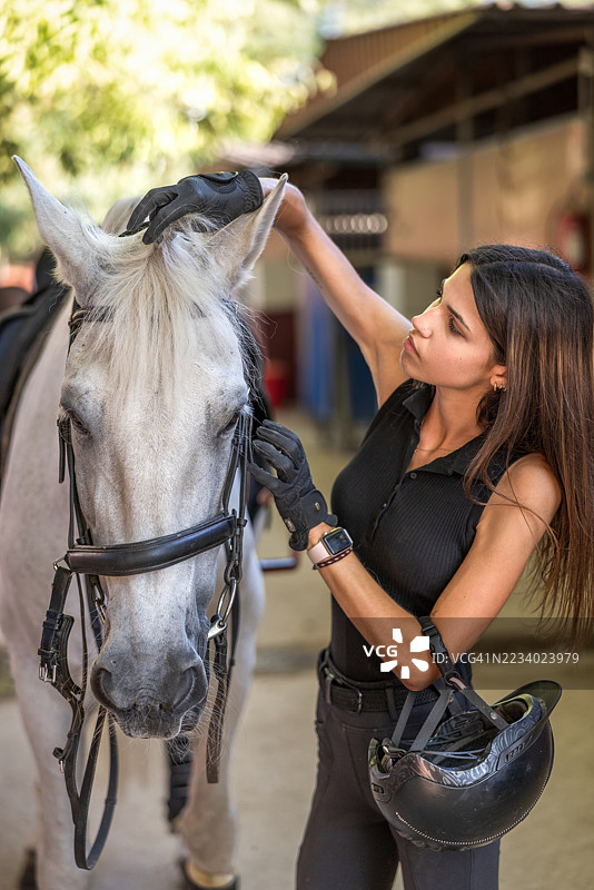 年轻的西班牙女骑手在日落时分为她的白马准备训练，调整马嚼子，展现出关爱与连接。图片素材