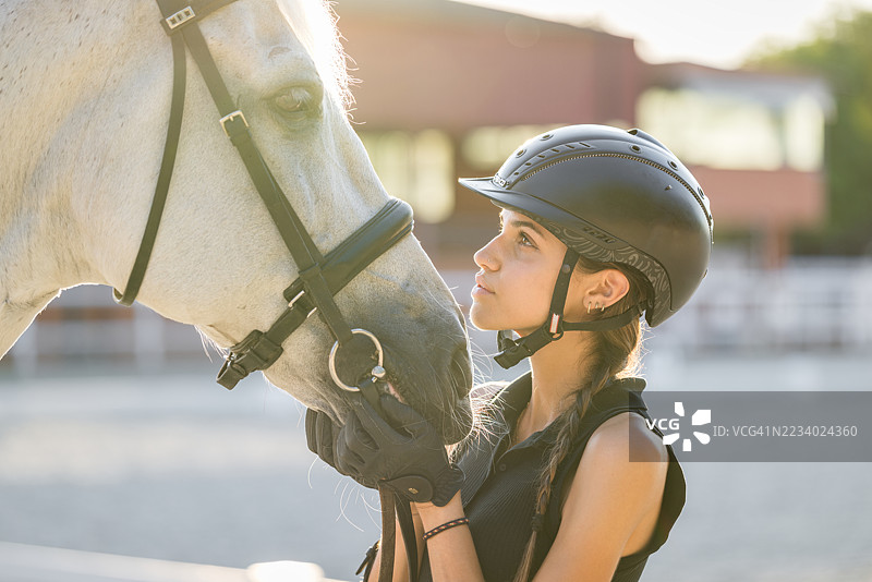 年轻的女骑手戴着手套和头盔，在骑马场的美丽日落中轻柔地抚摸着马的头，体现了骑手与动物之间的深厚纽带。图片素材