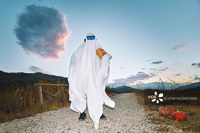 穿着白色床单的鬼魂装扮的人在秋季田野小路上手持南瓜。图片素材