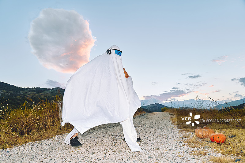 穿着白色床单的鬼魂装扮的人在秋季田野小路上手持南瓜。图片素材