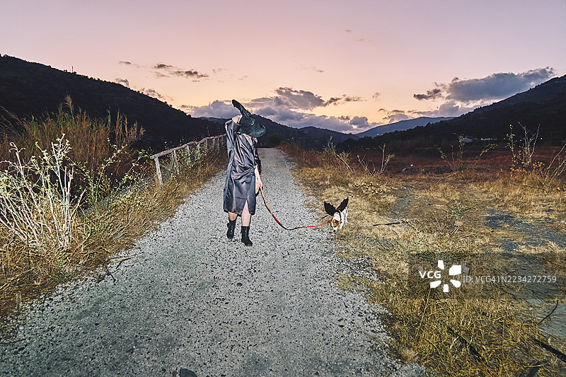 红发女性穿着女巫服装，带着她的杰克罗素梗狗在乡村夜晚散步，狗狗穿着蝙蝠翅膀的万圣节服装。图片素材