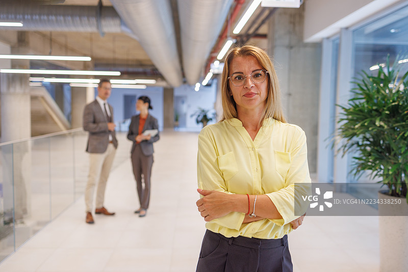 自信的女商人在现代办公室大堂站立，背景中同事们正在讨论图片素材