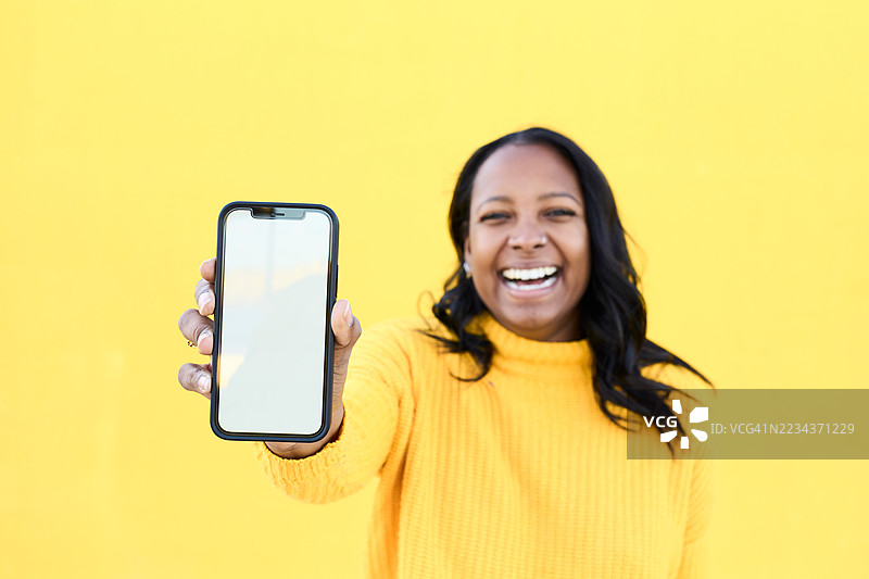 快乐的女性在黄色背景上展示带有空白屏幕的智能手机。图片素材