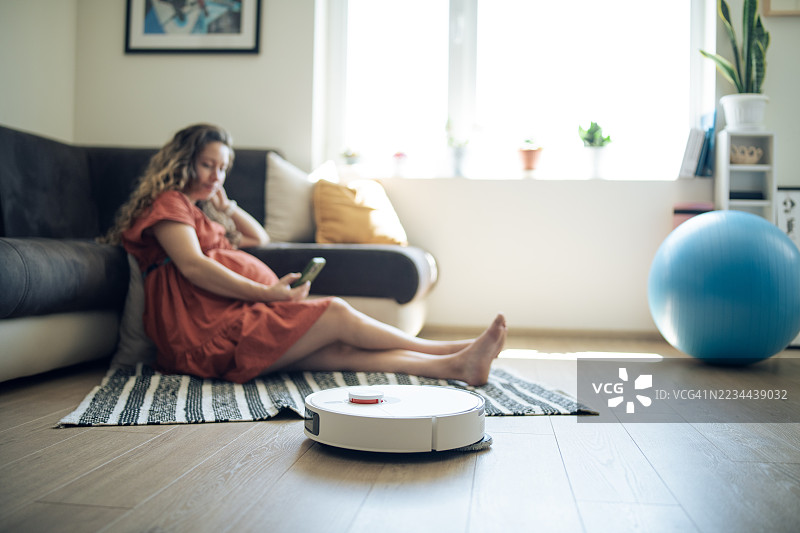 孕妇在休息，机器人吸尘器正在清洁地板。图片素材