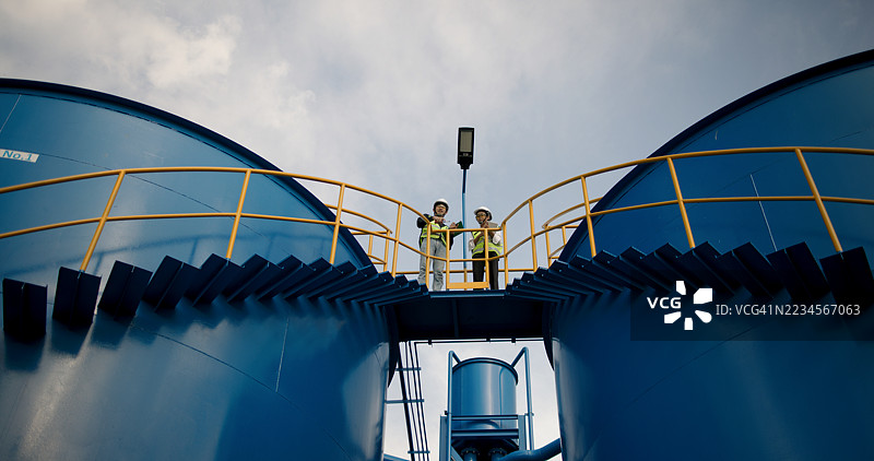 两名工程师穿着安全头盔和马甲，正走上连接到大型蓝色工业水箱的外部楼梯，进行水处理工作。图片素材