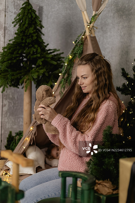 一个可爱的年轻女子抱着泰迪熊，坐在圣诞树旁，旁边还有一个小女孩的玩耍帐篷。图片素材