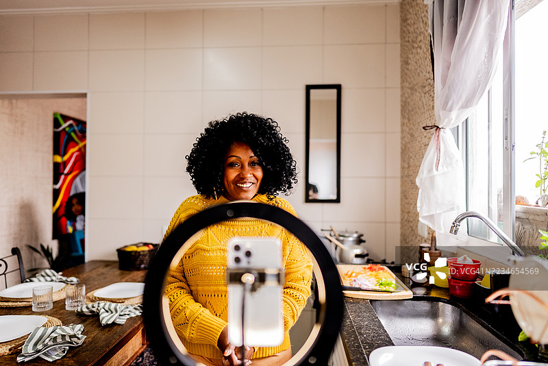 一位成熟的女性网红在家里的厨房用手机拍摄烹饪教程。图片素材