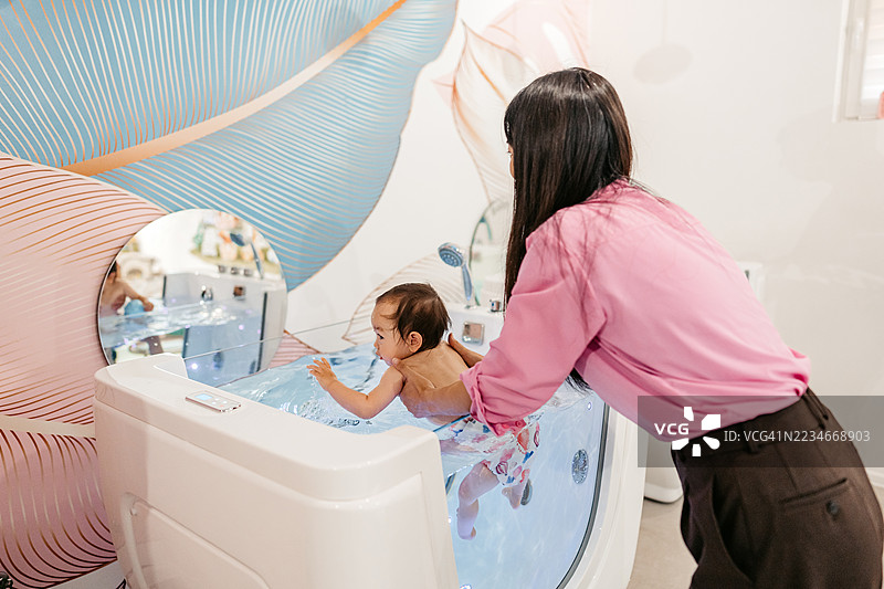 中年母亲抱着她的女婴，在婴儿水疗中心的浴缸里游泳，旁边有一个浮筒。图片素材