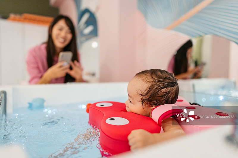 中年母亲用手机拍摄她的女婴在婴儿水疗中心的浴缸里游泳，旁边有浮筒。图片素材