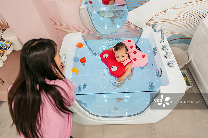 中年母亲在婴儿水疗中心看着她的女儿在浴缸里用浮具游泳图片素材