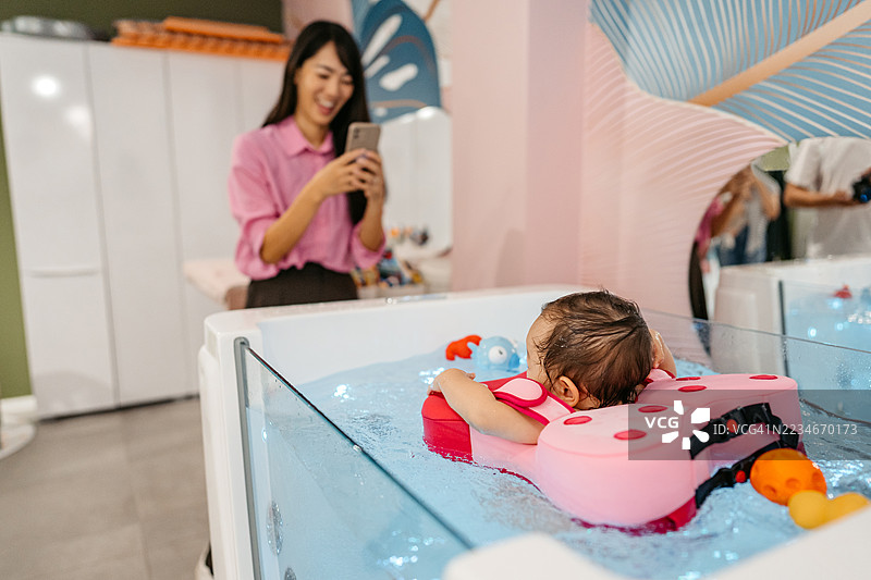 中年母亲用手机拍摄她的女儿在婴儿水疗中心的浴缸里游泳，女儿身边有一个浮筒。图片素材